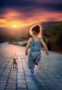 Kind mit hochgsteckten Haaren und in blau gemusterten Overall läuft auf einen Sonnenuntergang zu. Dem Kind entgegen kommt eine Jungtier einer Großkatze, es ist deutlich kleiner als das Kind.